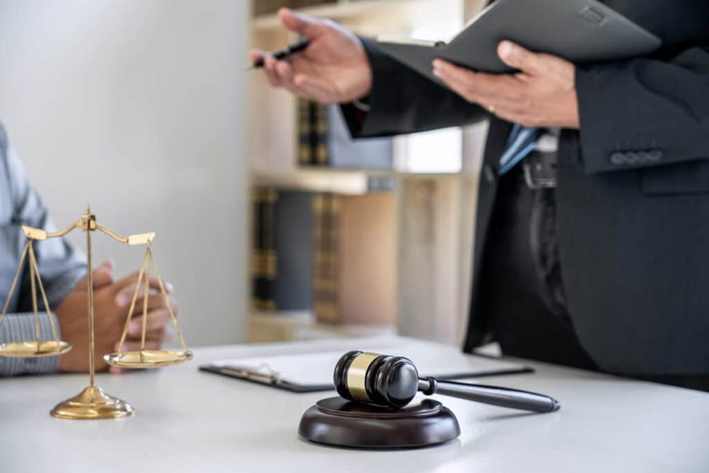 A catastrophic injury lawyer is standing at his desk, reviewing a case with a client. On his desk is paperwork, a gavel, and the scales of justice.