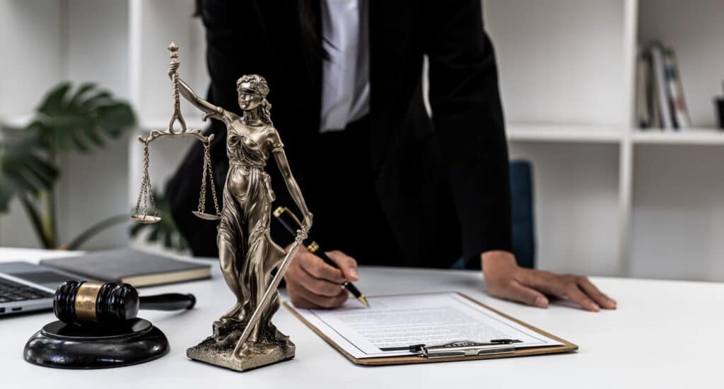 A catastrophic car accident injury attorney is working on paperwork at her desk. Next to her is a laptop, a Lady Justice statue, and a gavel.