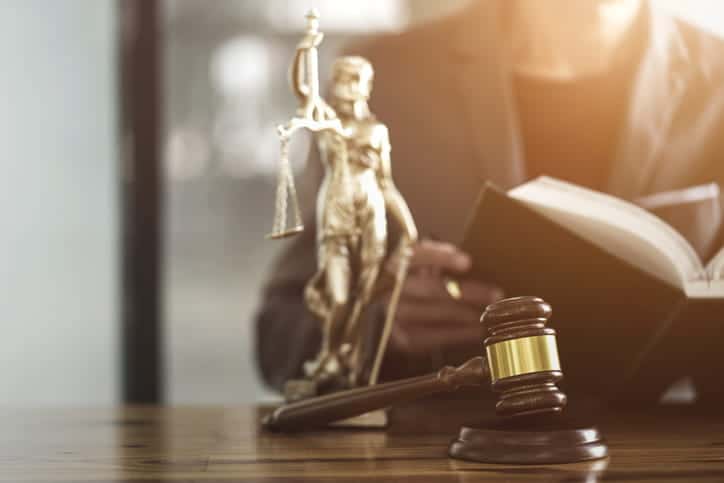 A Lady Justice statue and a gavel sitting on a desk as a personal injury attorney reads a book in the background. 