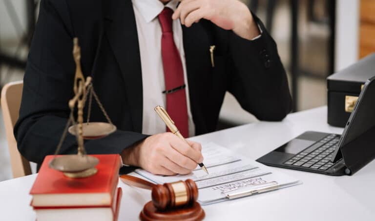 A catastrophic injury attorney at his desk filling out a contract agreement. Next to him is an open laptop, a gavel, and the scales of justice on top of a stack of books.